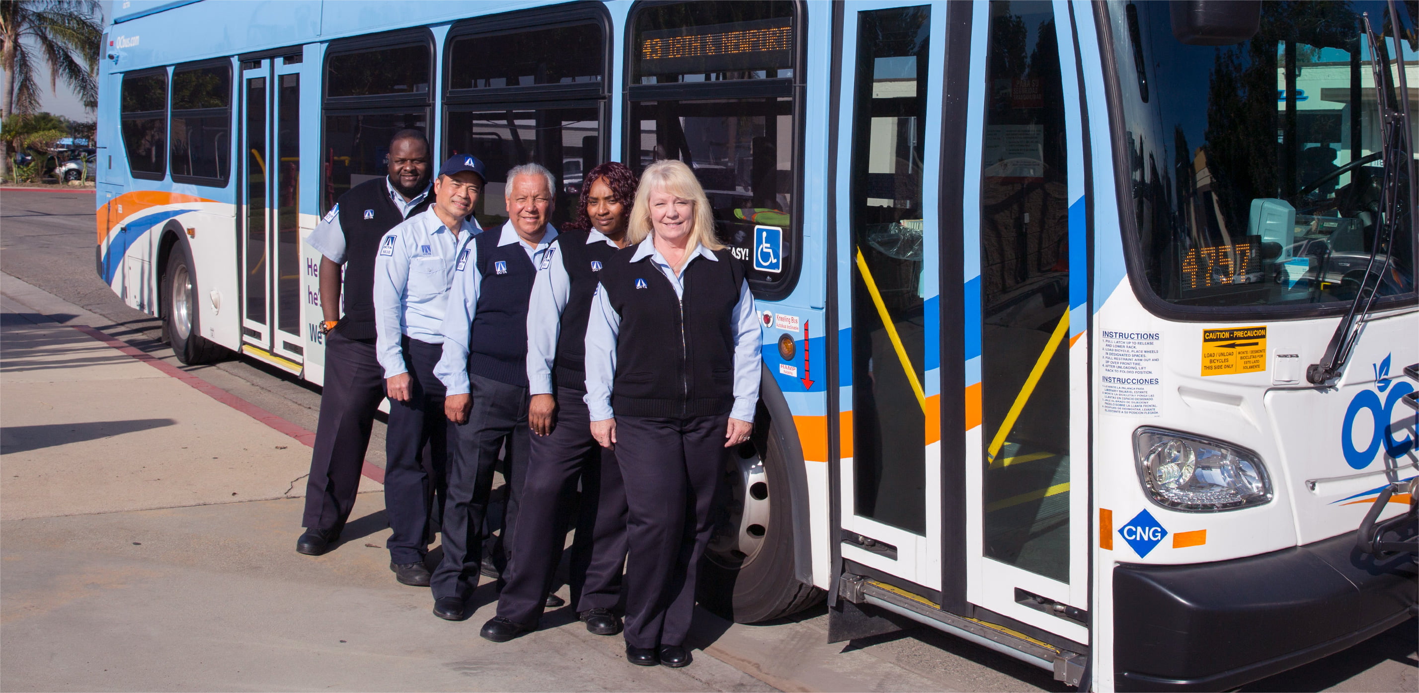 A group of smiling OC Bus drivers stand in a row in front of an OC Bus