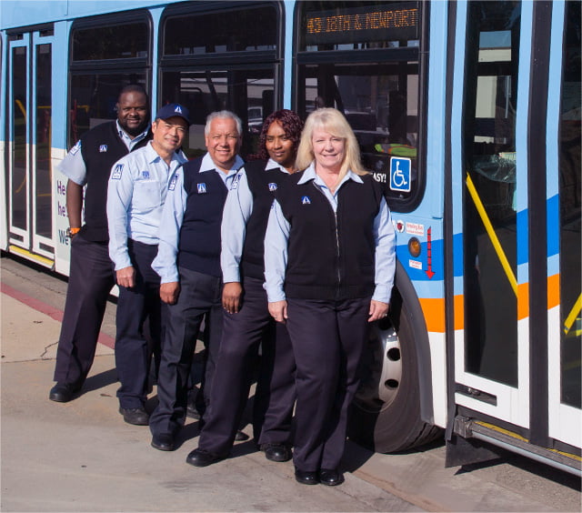 A group of smiling OC Bus drivers stand in a row in front of an OC Bus